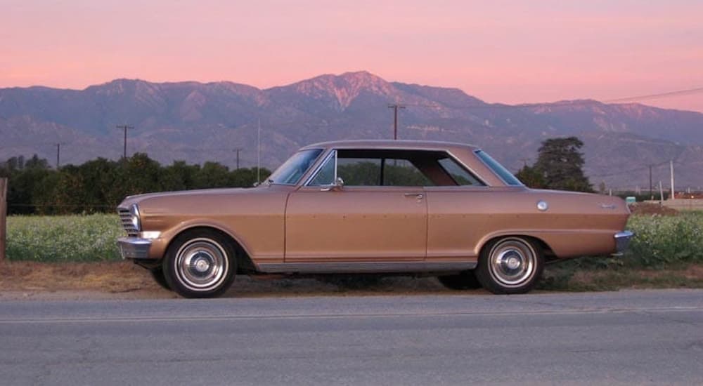 A tan 1863 Chevy Nova is parked in front of a pink sunset.