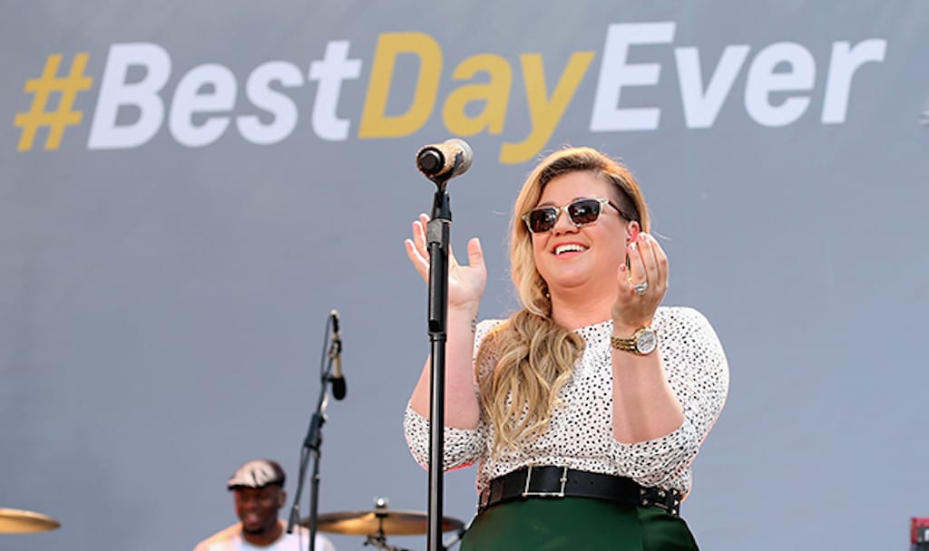 Kelly Clarkson is at the mic with a #BestDayEver behind her.