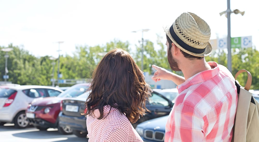 A man and a woman are pointing to a car in a parking lot outside.