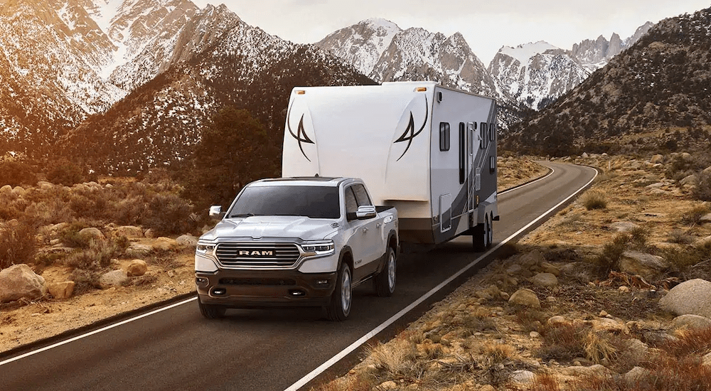 A white 2019 Ram 1500 is towing a camper with mountains in the distance. Check one out at a Ram dealership near you.