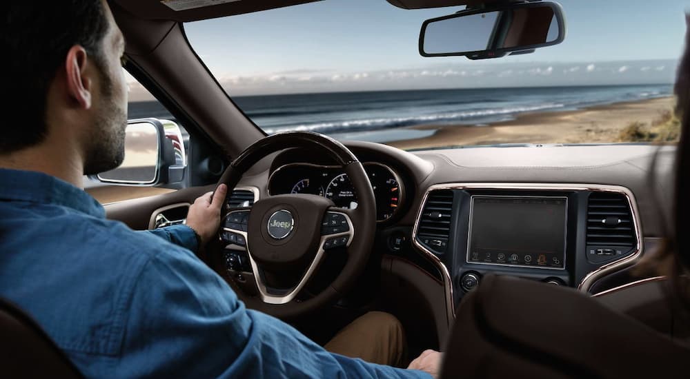The brown interior of a 2018 Jeep Grand Cherokee is shown with a male driver on the beach. Looking at a used Jeep Grand Cherokee is a great way to get into a like-new 2018.