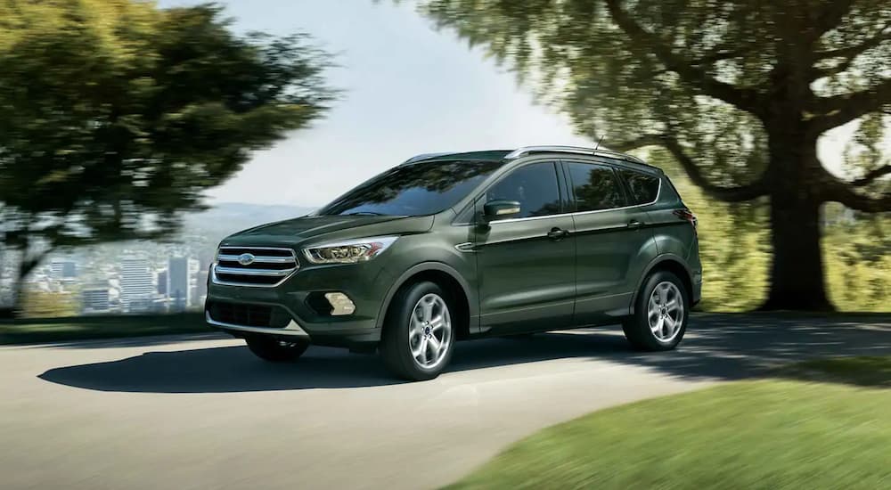 A green 2019 Ford Escape is driving on a narrow road with trees and a city view behind it. Check out performance when comparing the 2019 Ford Escape vs 2019 Chevy Equinox.