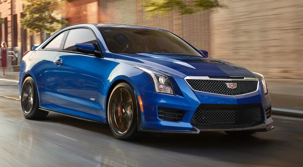 A blue 2019 Cadillac ATS-V, available among older model Cadillac used cars as well, is driving in front of brick buildings. driving in front of buildings.