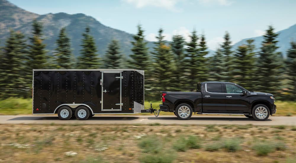 A black 2019 Chevy Silverado 1500 is shown towing a large trailer with trees and mountains in the background. 