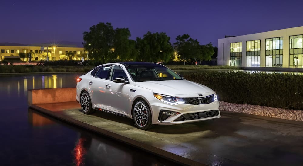 A white Kia Optima is parked on a platform with trees and lights in the background, and could be found with Kia lease deals. 