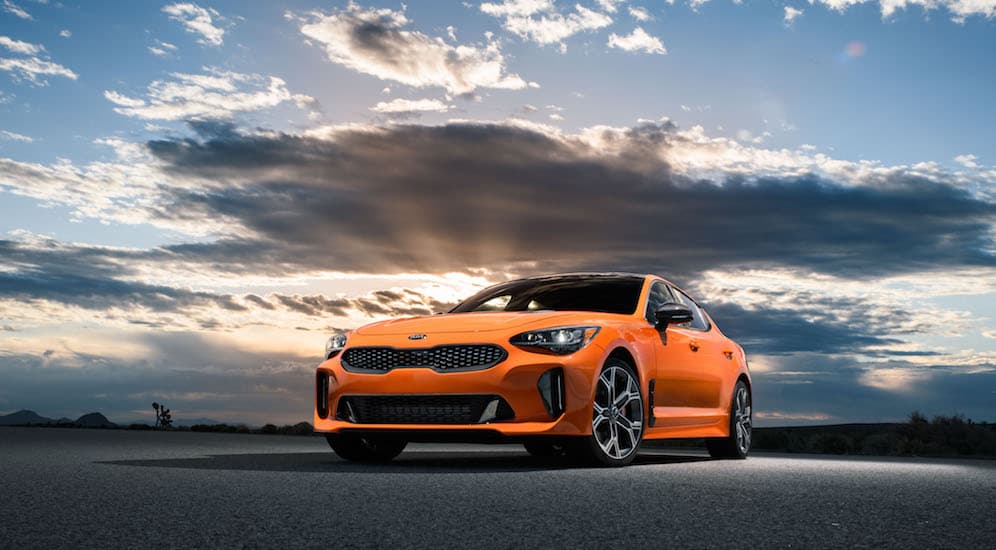 A 2020 orange KIA Stinger GTS is parked in the parking lot with the sun in the background and could be found at a Kia dealer with lease deals..