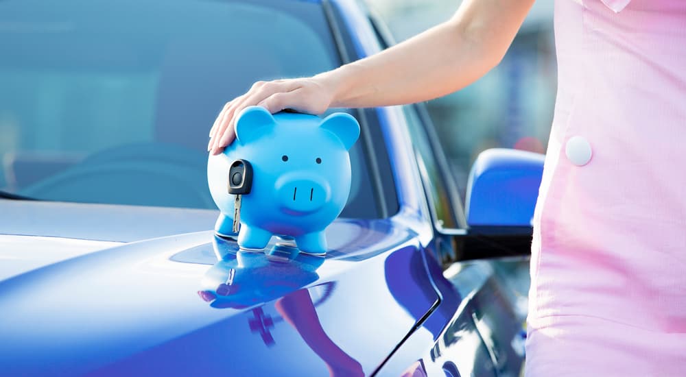 A closeup of a woman holding a piggy bank with car keys on the hood of a blue car is shown.