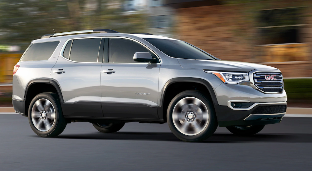 A silver 2019 GMC Acadia, which wins when comparing performance between the 2019 GMC Acadia vs Toyota Highlander, is driving past a tan building.