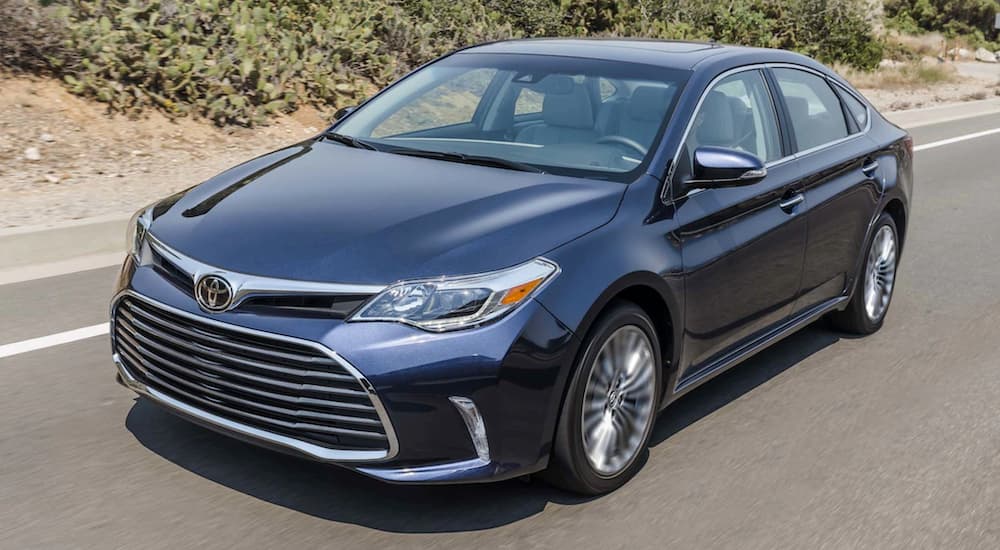 A blue 2017 Toyota Avalon, popular among used cars for sale, is driving with desert shrubs behind it.