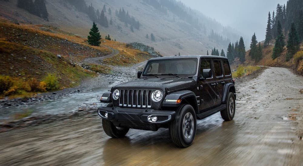 A black 2018 Jeep Wrangler is driving on a dirt road in front of a river and mountains.