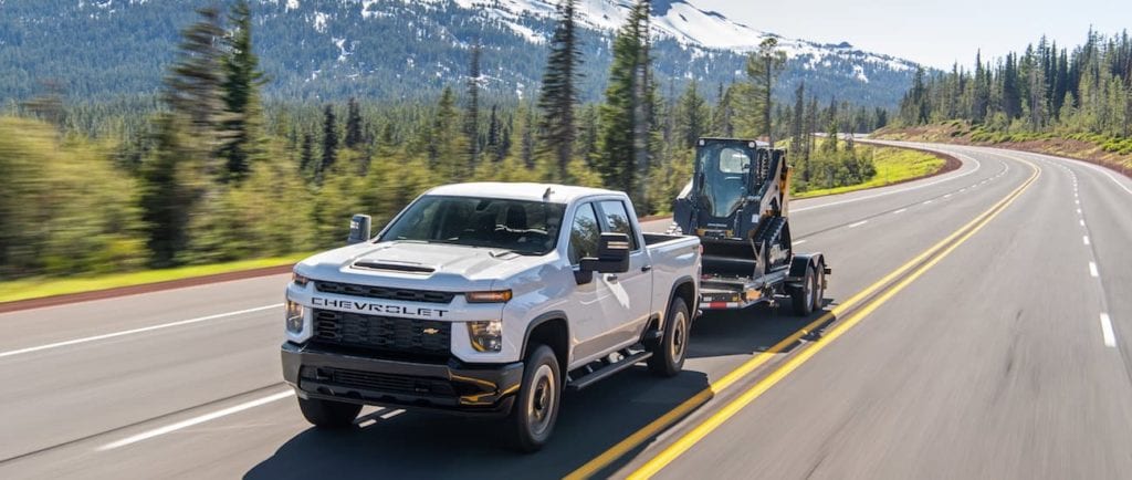 A white 2020 Chevy Silverado 2500HD is towing heavy equipment with mountains in the background. 