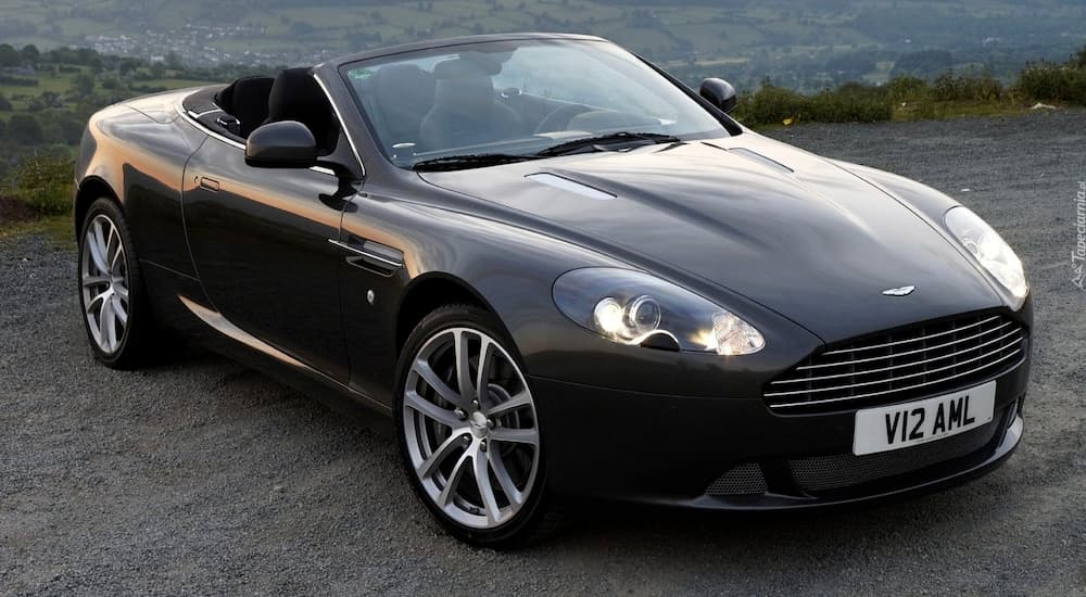 A black Aston Martin DB9 Volante is parked with mountains in the distance.