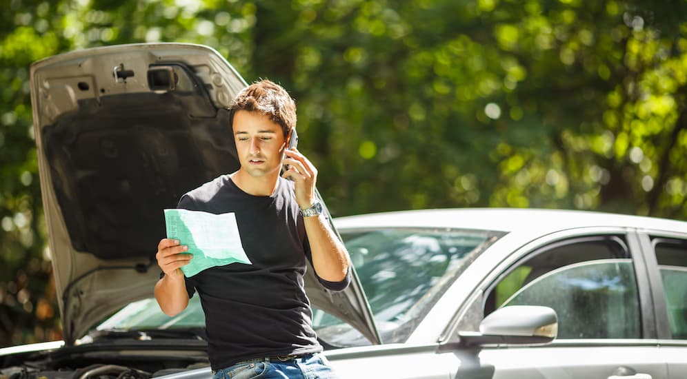 A man is standing next to his broken down used car  while looking at a paper and on the phone. 