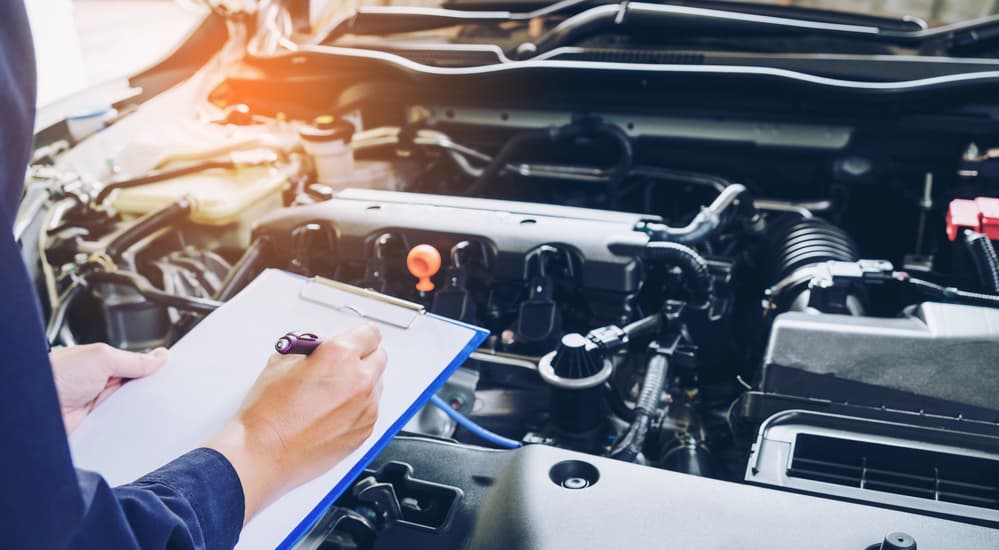 A mechanic is looking at a car engine while doing a quick lube service with a check list in his hand. 