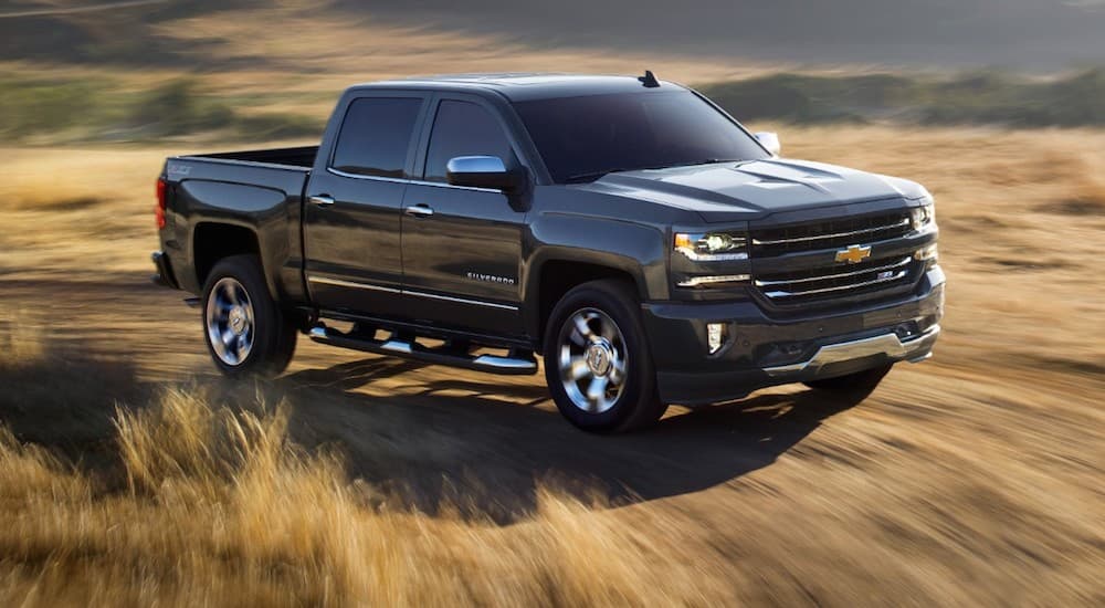 A dark grey 2018 Chevy Silverado is driving on a dirt backroad through a hay field. 