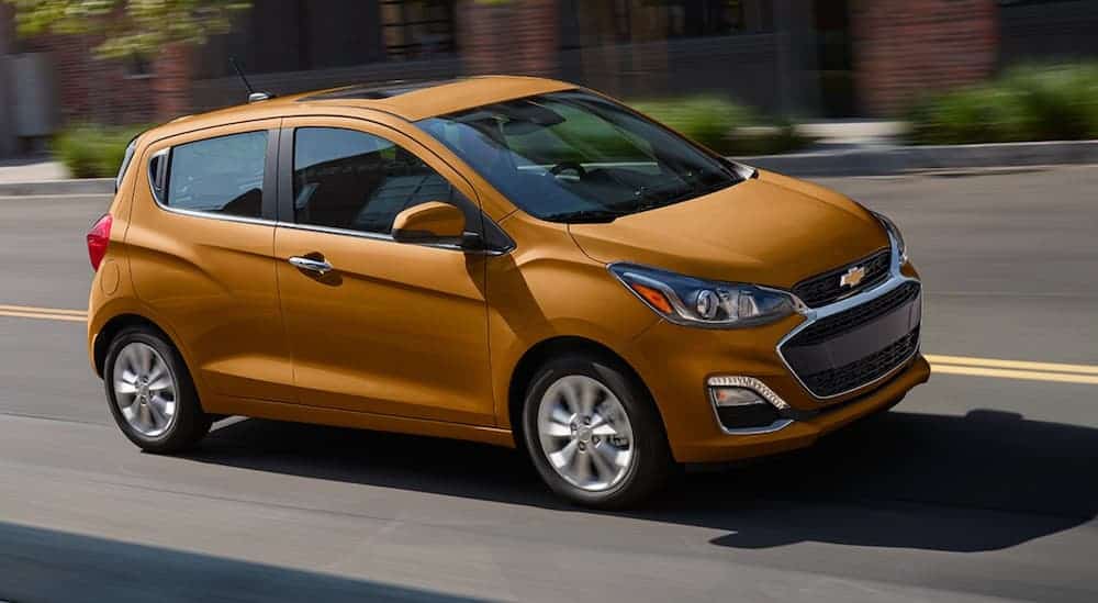 A gold colored 2020 Chevy Spark is driving on an empty city street during the day. 