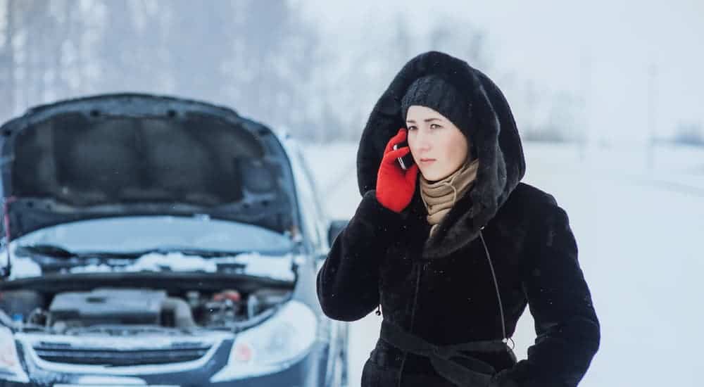 A woman is on the phone with a broken down car behind her in the snow.