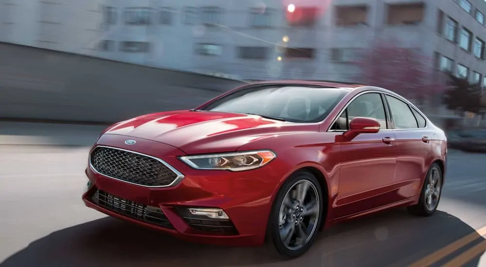 A red 2019 Ford Fusion, which wins when comparing the 2019 Ford Fusion vs 2019 Toyota Camry, is driving on a city street on a sunny day.