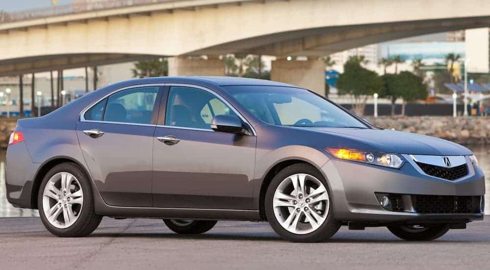 A grey 2010 Acura TSX is parked in a empty parking lot next to a bridge. 