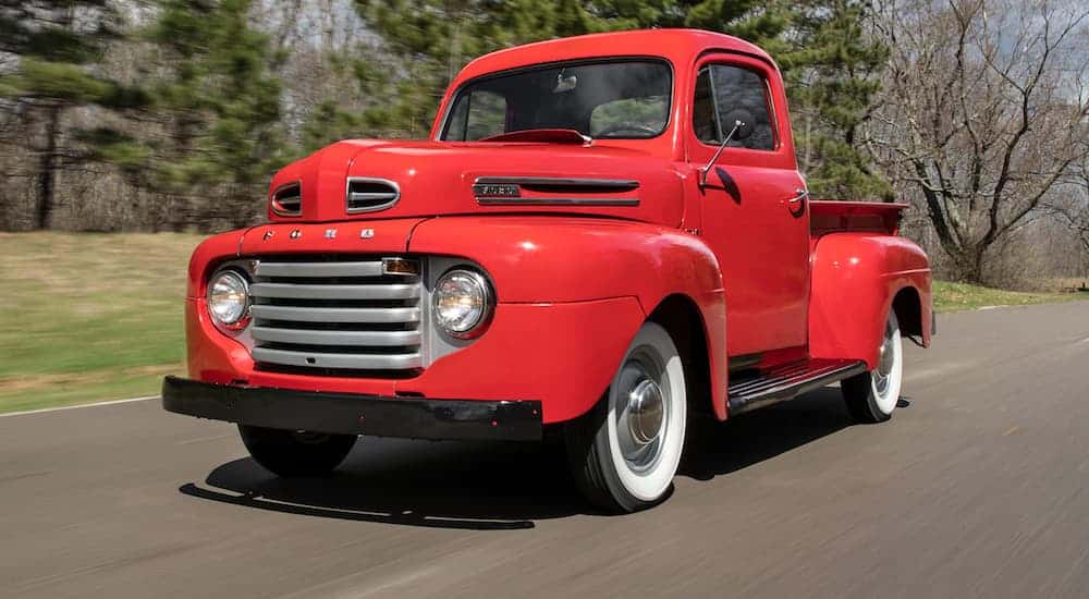 A red 1949 Ford F-1 is driving on a treelined road on a sunny day. 