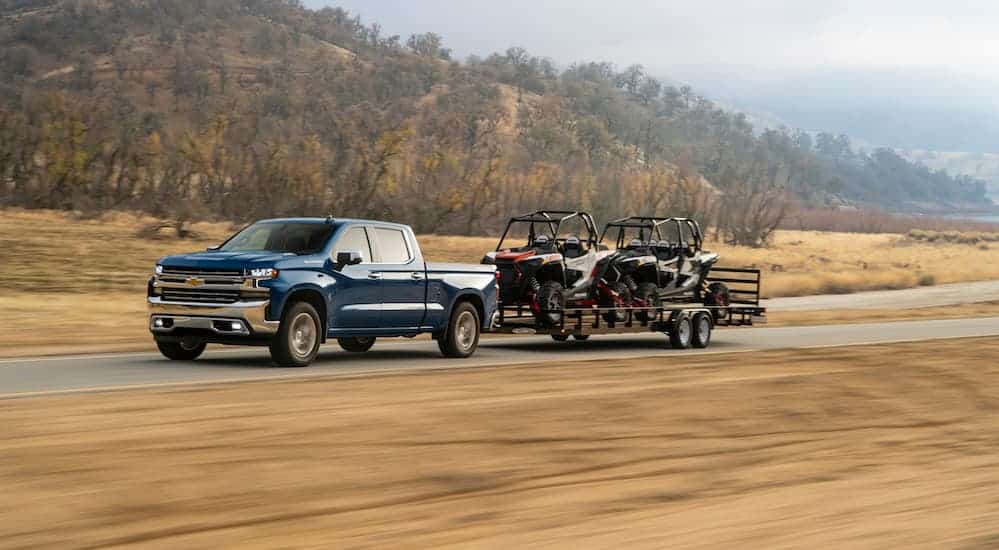 A blue 2020 Chevy Silverado 1500 diesel is towing 2 side by sides on a trailer past a grassy field. 
