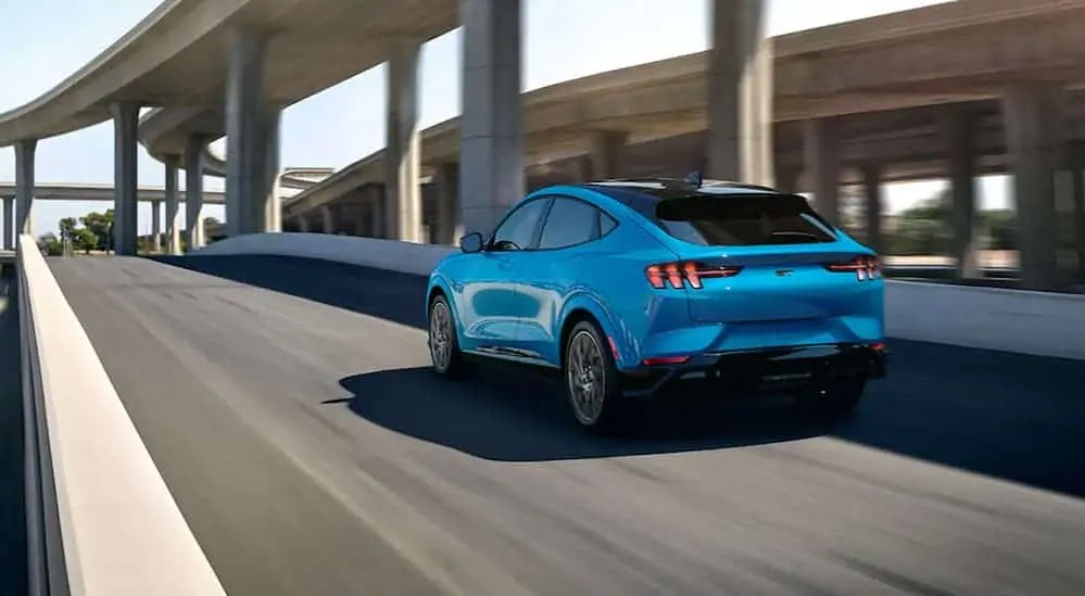 A blue 2021 Ford Mustang Mach-E GT is driving through a city under a highway bridge.