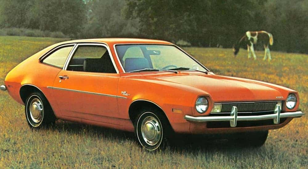 An orange Ford Pinto, which is known to be one of the worst used cars on the market, is parked in a grassy field with a horse in the background. 
