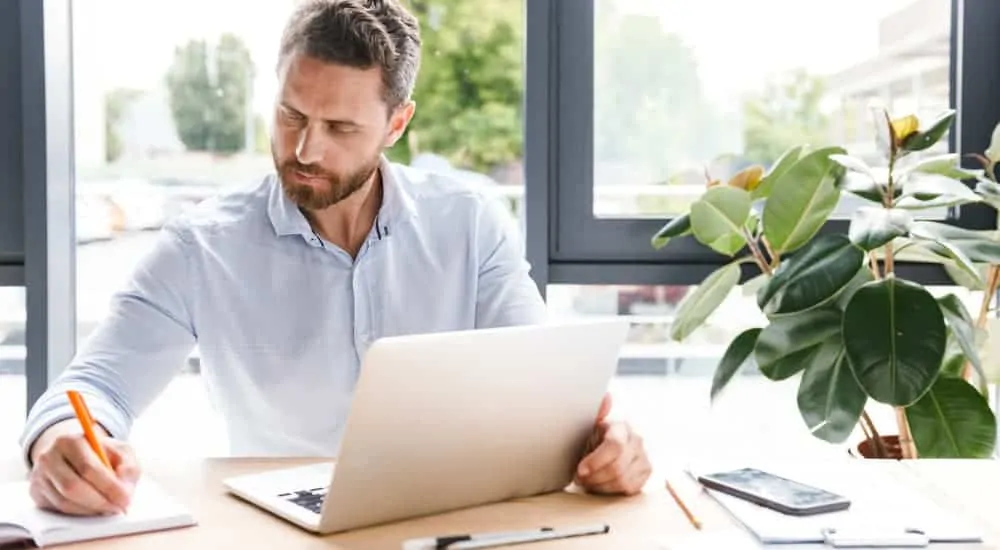 A man is researching used cars for sale on his laptop and taking notes.