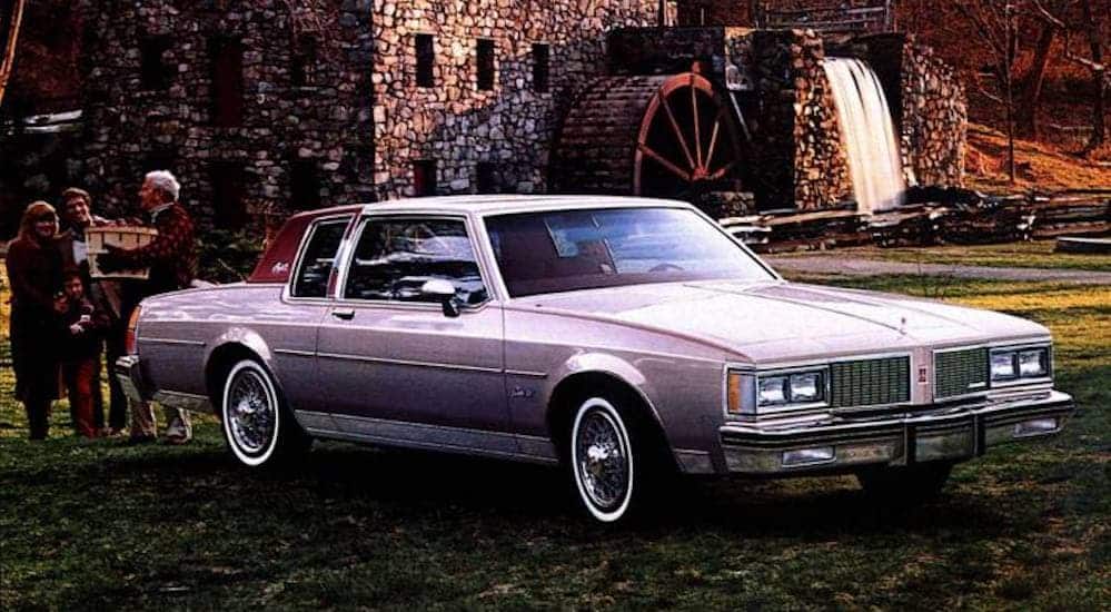 A light purple 1979 Oldsmobile Diesel is parked in front of an old stone building on a cobble stone driveway.
