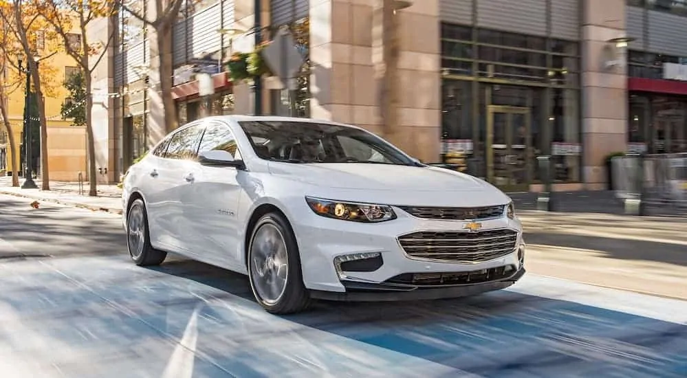 A white 2018 Chevy Malibu, which is a popular model among used cars near me, is driving past an outdoor restaurant. 