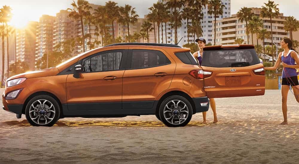 Two friends are standing next to their orange 2020 Ford Ecosport that's parked on the beach during sunset.