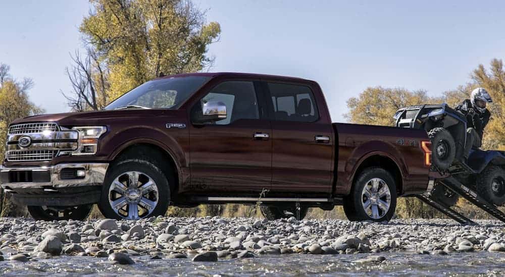 A side view of a red 2020 Ford F-150, which wins when comparing the 2020 Ford F-150 vs 2020 Chevy Silverado, is parked on gravel while a man loads his ATV into the truck bed. 