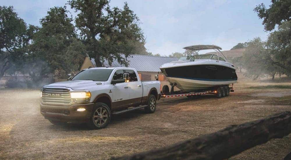 A white 2020 Ram 2500 with a Cummins diesel engine is towing a large boat in front of a farm at dusk. 