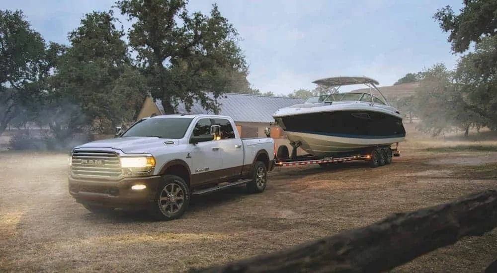 A white 2020 Ram 2500 with a Cummins diesel engine is towing a large boat in front of a farm at dusk. 