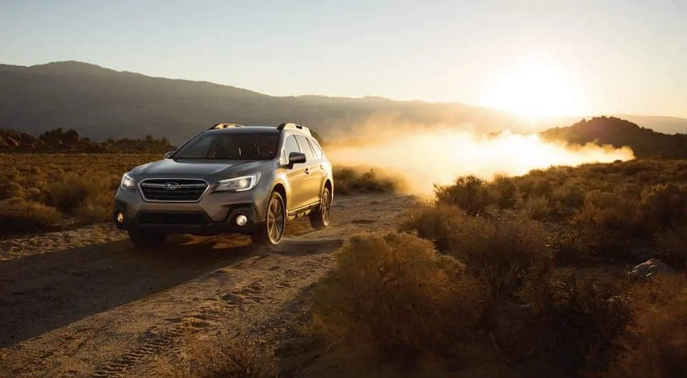 A silver 2018 Subaru Outback is driving on a dirt road at sunset.