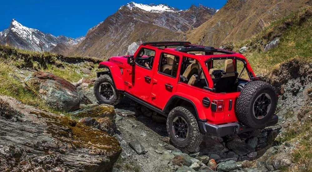 A red 4-door 2020 Jeep Wrangler, which is a top Jeep Wrangler for sale, is rock crawling up a hill with mountains in the distance. 