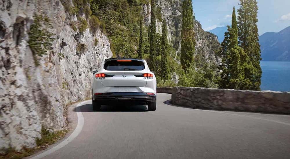 A white 2021 Ford Mustang Mach-E, which wins when comparing the 2021 Ford Mustang Mach-E vs Tesla Model Y, is driving on a curvy road next to a lake. 