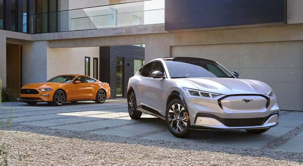 A silver 2021 Ford Mustang Mach-E, which is a soon to be popular option among Ford SUVs, is parked next to an orange 2020 Ford Mustang in front of a home. 