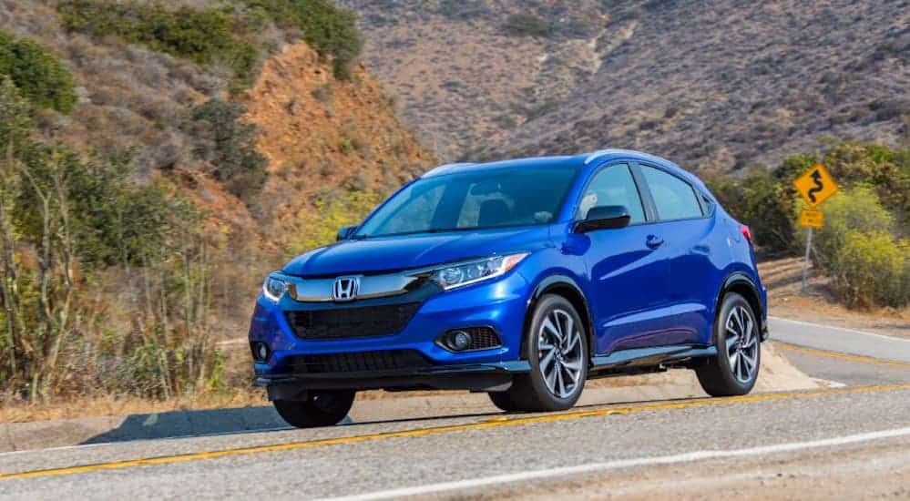 A blue 2020 Honda HR-V is driving around a treelined corner. 