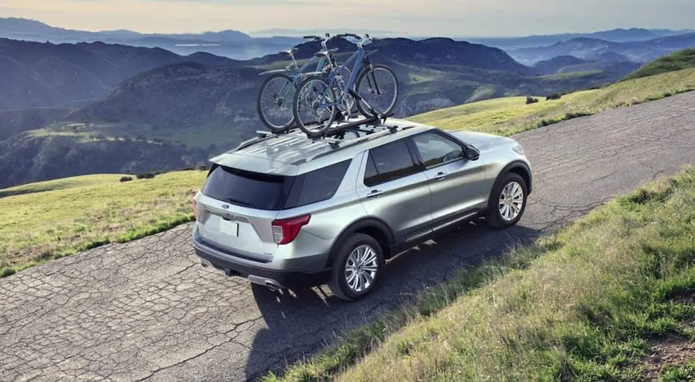 A silver 2020 Ford Explorer is driving on a dirt road overlooking mountains with bikes on the roof rack.
