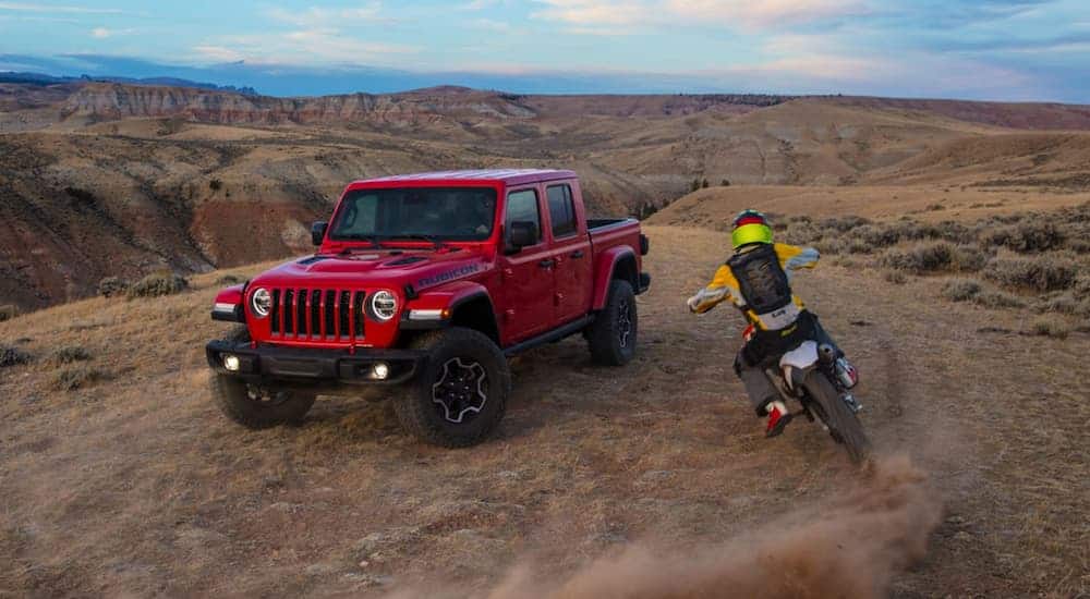 A dirt biker is riding around a red 2020 Jeep Gladiator Rubicon in the desert.