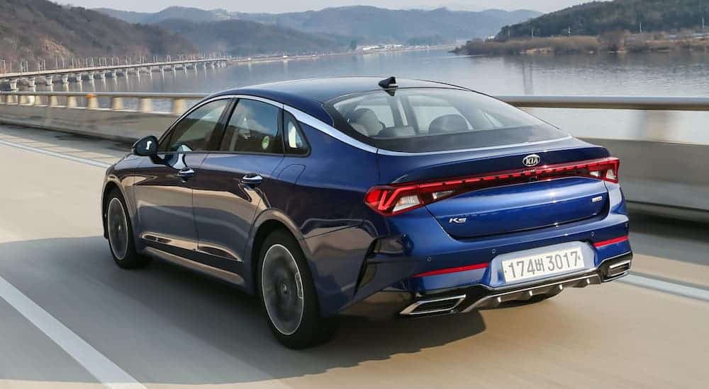 A blue 2021 Kia Optima is driving on highway that's next to a lake.