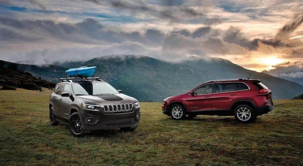 A gray 2020 Jeep Cherokee with a kayak next to a red Cherokee atop a mountain with a vibrant sunset behind distant mountains