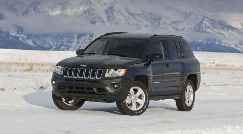 A black 2011 Jeep Compass is parked on snow in front of mountains.
