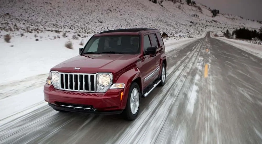 A red 2011 Jeep Patriot is driving on a snowy road after leaving a used car dealership.