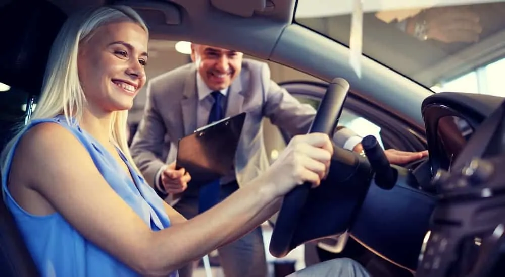 A woman is in a used car while a salesman shows her features.