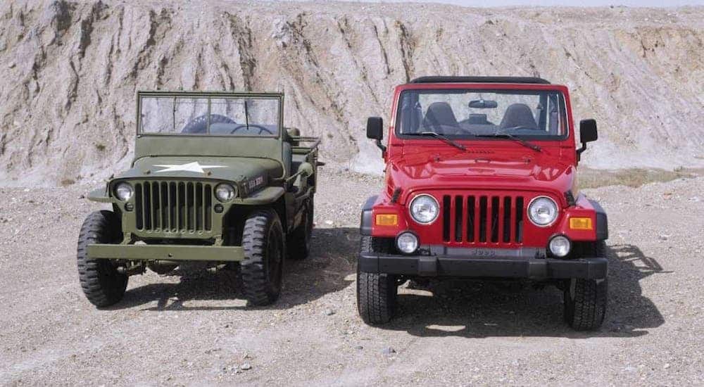 A 1940s Jeep is parked next to a red early 2000s Wrangler in front of a sand pile.