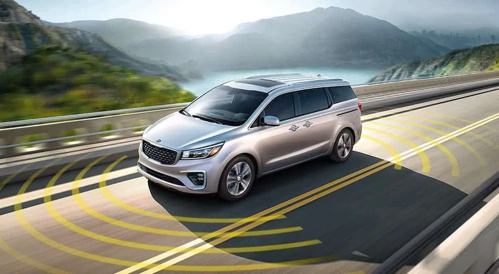 A silver 2020 Kia Sedona is driving on a highway with simulated safety monitoring graphics.