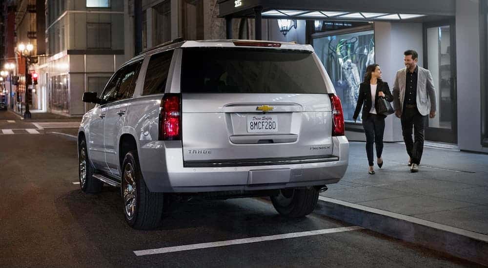 A silver 2020 Chevy Tahoe is parked on a city street at night with a couple walking toward it.