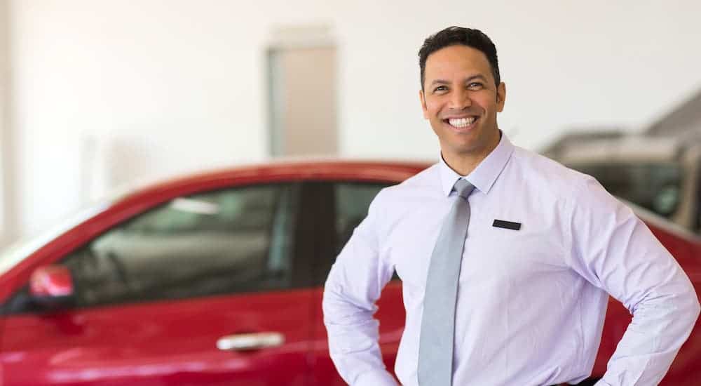 A smiling salesman is standing in front of a red car in a at a Chevy dealership showroom.